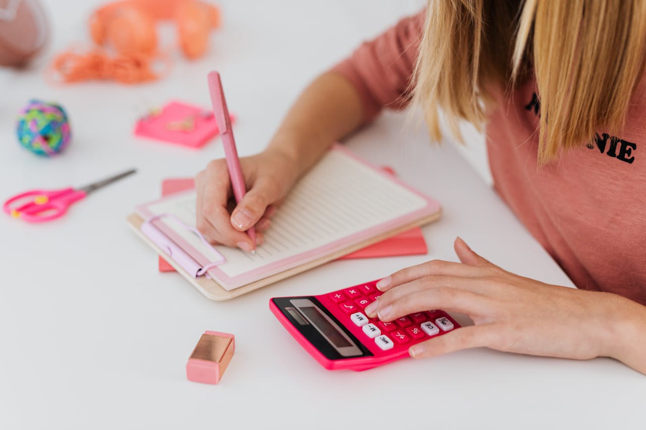 about-us A young student uses a pink calculator while writing notes in a notebook, surrounded by colorful stationery.