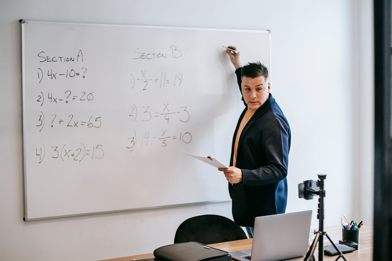 A teacher explains math problems on a whiteboard in a modern classroom setting.