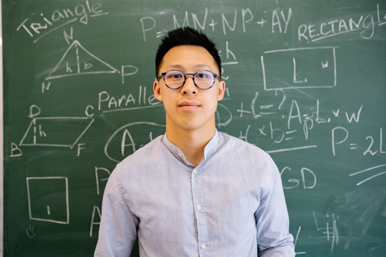 heros-img Educator standing before chalkboard filled with geometry and algebra equations, wearing glasses and long-sleeved shirt.